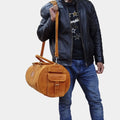 A guy in black jacket and blue jeans hanging costal travel duffle on shoulder posing