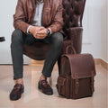 Urban Nomad Leather Backpack placed besides model in chair with green pants 