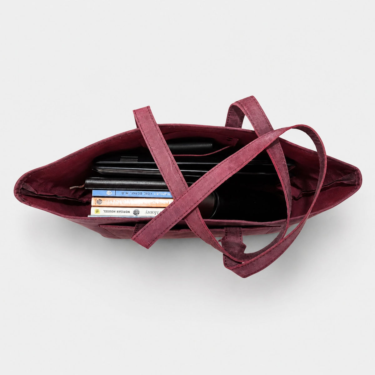 Top view looking inside a burgundy leather laptop tote bag packed with a black laptop, notebooks, water bottle, and accessories