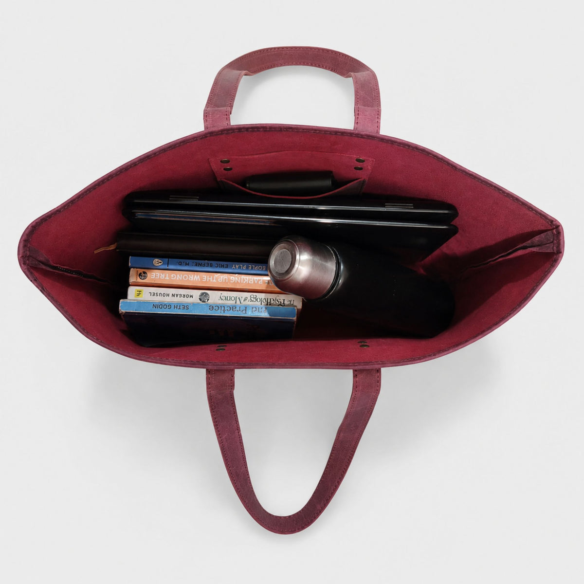Overhead view looking inside an open burgundy leather work tote bag showing inner pocket storage, a black laptop, books, and a stainless steel water bottle