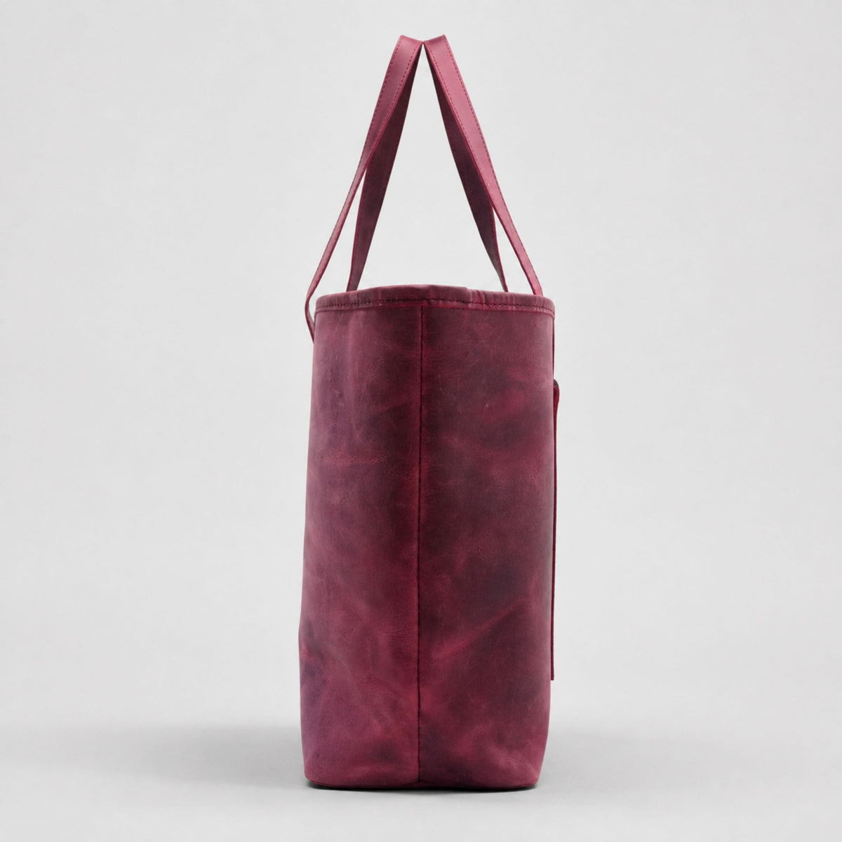 Side view profile of a structured burgundy leather shoulder tote bag highlighting the clean stitched seams, wide bottom base, and flexible shoulder straps